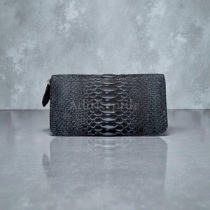 May include: A dark gray wallet with a textured, snake-like pattern. The rectangular wallet has a zipper closure on one side and a smooth, black zipper pull. The wallet is displayed on a reflective surface.