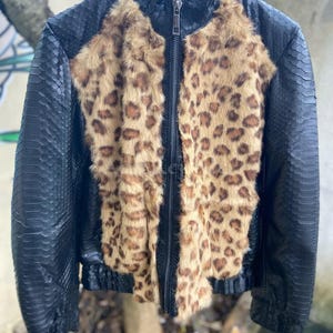 Könnte beinhalten: Eine schwarze Jacke mit einer strukturierten schlangenlederartigen Optik und einem vertikalen Einsatz aus braun-schwarzem Leopardenfell auf beiden Seiten des Frontreißverschlusses. Das Kleidungsstück hat einen hohen Kragen und lange Ärmel.