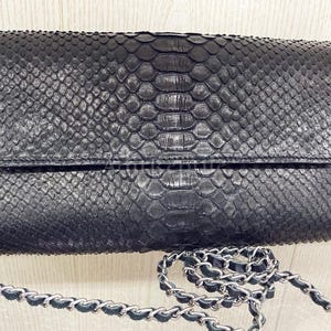 May include: A black clutch with a snakeskin pattern and a silver chain strap. The rectangular bag has a flap closure and a textured surface. The chain is made of silver metal links and black leather.