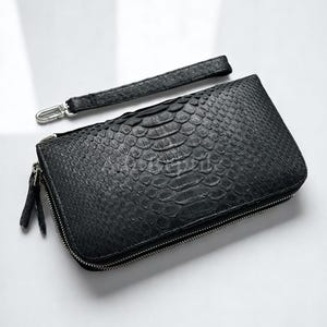 Puede incluir: Cartera de cuero negro con textura de piel de reptil, cierre de cremallera y correa de muñeca desmontable. La cartera es rectangular y tiene una cremallera plateada. La correa de muñeca mide unos 20 cm.
