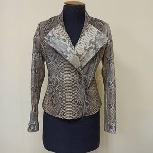 May include: A snakeskin-patterned jacket with a diagonal zipper closure. The jacket features a wide collar and long sleeves. The color palette includes shades of brown, gray, and beige, creating a textured appearance. The jacket is displayed on a mannequin.