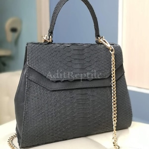 May include: A black handbag with a textured snakeskin pattern. The bag has a top handle and a gold chain strap. The flap closure is detailed with a geometric design. The text "AditReptile" is visible on the front of the bag.