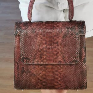 Puede incluir: Bolso cuadrado con solapa, hecho de un material similar a la piel de serpiente en tonos marrón y burdeos. El bolso tiene un asa superior y una superficie texturizada, con el texto "AdieReptile" visible en la parte delantera.