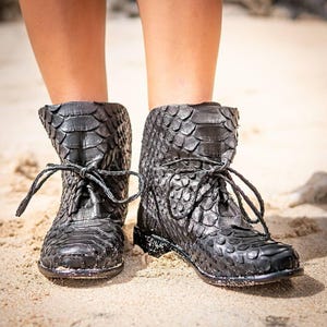 Könnte beinhalten: Schwarze Stiefeletten mit einem strukturierten, schuppenartigen Muster und Schnürverschlüssen. Die Stiefel werden an einem Sandstrand getragen. Die Stiefel scheinen aus Leder oder einem ähnlichen Material zu sein.