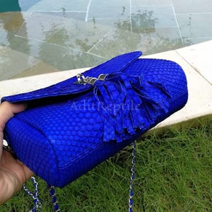 May include: A vibrant blue handbag with a textured, scaly pattern. The bag features a flap closure, a silver chain strap, and a decorative tassel. The brand name "AditReptile" is visible on the bag.