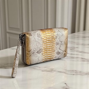 May include: A rectangular clutch bag with a snakeskin pattern in white, gold, and grey. It features a zip closure and a wrist strap. The accessory is on a marble surface.