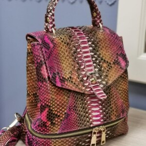 May include: A multi-colored backpack with a handle and a flap closure. The bag features a snakeskin pattern in shades of pink, brown, and gold. It has a zipper closure and a small handle at the top. The bag is approximately 12 inches tall.