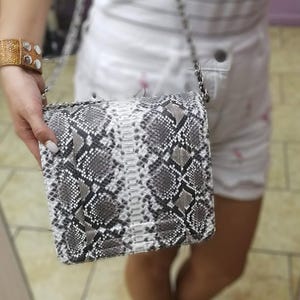 May include: A gray, white, and black snakeskin-patterned handbag with a silver chain strap. The bag is rectangular with a flap closure. A gold-toned bracelet with white stones is visible on the left wrist.