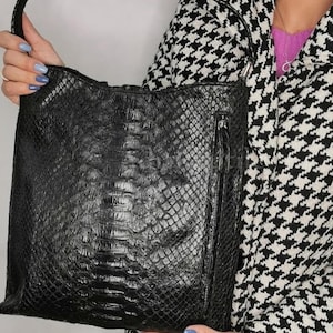 May include: A black leather shoulder bag with a textured, reptile-like pattern. The bag has a zipper closure and a braided handle. The background includes a black and white houndstooth pattern.