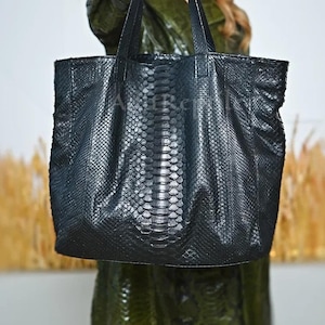 May include: A black tote bag with a snakeskin pattern. The bag has two handles and a rectangular shape. The bag is made of a textured material and appears to be leather. The bag is held up by a person.