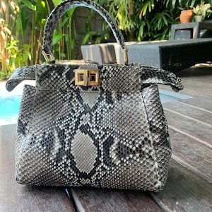 May include: A grey and black snakeskin handbag with a top handle and a gold-coloured clasp. The bag has a structured shape and a flap closure. The background includes greenery and a wooden surface.