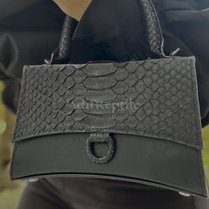 May include: Black handbag with a textured snakeskin pattern on the top flap and handle. The bottom portion of the bag is smooth black leather. The bag has a metal ring detail and the text "Adit Reptile".
