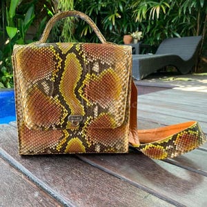 May include: A square-shaped handbag with a handle and a shoulder strap. The bag is made of snakeskin with a pattern of brown, yellow, and tan scales. The bag has a metal clasp closure.