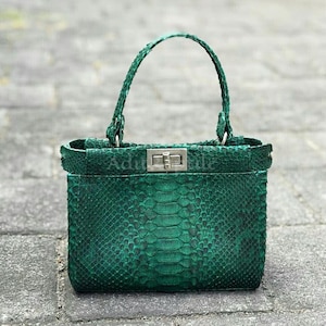 May include: A green handbag with a textured snakeskin pattern. The bag has a top handle and a silver-colored clasp. The bag is rectangular with a structured design. The handle is braided and attached to the bag with silver hardware.