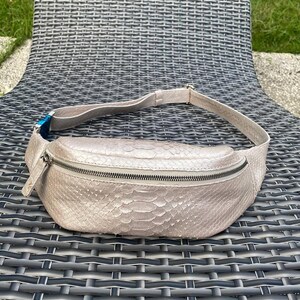 May include: A light beige fanny pack with a snakeskin pattern. The bag has a zipper closure and an adjustable strap. The fanny pack is made of a textured material and is designed to be worn around the waist.