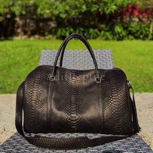 May include: A black leather duffel bag with a snakeskin pattern. The bag has two handles and a shoulder strap. The bag is sitting on a gray wicker chair. The background is a green lawn and trees.