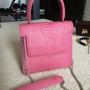 May include: A pink handbag with a textured snakeskin pattern. The bag has a top handle and a detachable chain strap. The bag has a flap closure and a silver chain strap. The bag is approximately 8 inches tall.