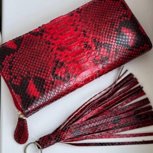 Puede incluir: Una cartera roja y negra con textura de piel de serpiente y un llavero con borla a juego. La cartera tiene un cierre de cremallera con un tirador dorado. El llavero tiene un anillo plateado.