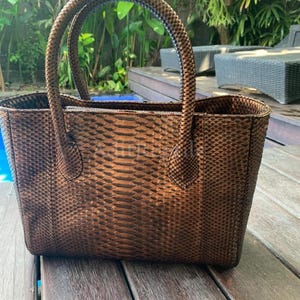 May include: A brown tote bag with a snakeskin pattern. The bag has two rounded handles and a rectangular shape. The bag is sitting on a wooden surface.