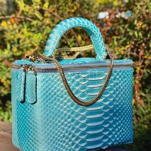 May include: A turquoise, rectangular handbag with a textured, snakeskin-like pattern. The bag features a matching handle, a gold-colored chain strap, and a gold zipper closure. The bag is sitting on a wooden surface.