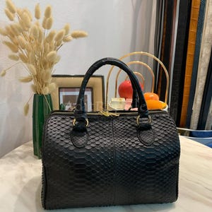 May include: A black leather handbag with a textured, scale-like pattern. The bag has two handles, a zipper closure, and gold-colored hardware. The bag is sitting on a marble-topped table.