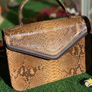 May include: A brown and black snakeskin handbag with a top handle and a flap closure. The bag has a rectangular shape with a dark brown trim around the flap. The snakeskin pattern is visible on the exterior of the bag.