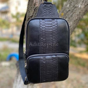 May include: Black leather sling bag with a central panel of snakeskin texture. The bag has a zippered main compartment and a smaller front pocket. The text "AditReptile" is visible on the front.