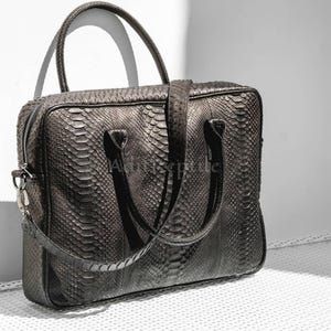 May include: A dark gray, rectangular briefcase with a textured snakeskin pattern. The bag has a top handle, a detachable shoulder strap, and a zipper closure. The bag is made of a durable material and is suitable for carrying documents and other essentials.