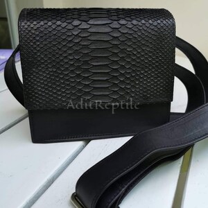 May include: Black crossbody bag with a textured, snake-like pattern on the flap. The bag has a rectangular shape with a solid black base and a long, adjustable strap. The text "AditReptile" is visible on the front.
