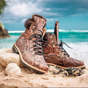 Könnte beinhalten: Ein Paar braune und beige Stiefel mit Schlangenmuster, schwarzen Schnürsenkeln und Fellbesatz. Die Stiefel stehen an einem Sandstrand mit dem Meer im Hintergrund. Robustes Design mit Metallösen.