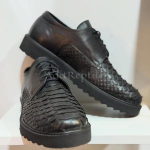 May include: A pair of black lace-up shoes featuring a textured, scale-like pattern on the front and sides, smooth leather panels at the heel and lace area, and a thick, black serrated rubber sole. The text AditReptile is visible on the side.