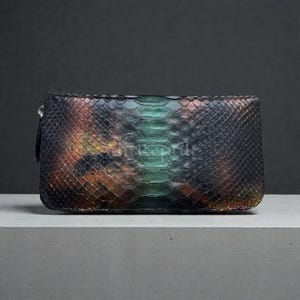 Puede incluir: Un bolso de mano rectangular con un estampado de piel de serpiente. La paleta de colores incluye tonos de verde, marrón y negro. El bolso de mano tiene una correa de muñeca en el lado izquierdo.