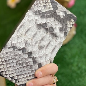 Puede incluir: Primer plano de una cartera con estampado de piel de serpiente en tonos grises y blancos. La cartera está sujeta con una mano que lleva un anillo dorado. Las escamas de la piel de serpiente son visibles.