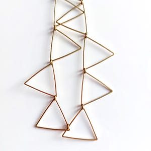 Puede incluir: Un collar dorado con un diseño geométrico de triángulos interconectados. Los triángulos están dispuestos en un patrón en cascada, creando una estética moderna y minimalista. El collar se muestra sobre un fondo blanco.