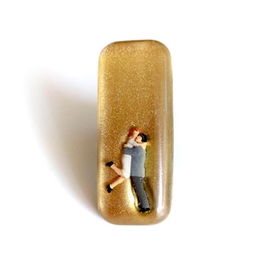 May include: A gold-coloured resin pendant in a rectangular shape. It features a miniature couple embracing. The man wears a grey suit, and the woman wears a white dress. The pendant is set against a white background.