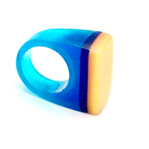 Op de afbeelding: Een dikke harsring met een kleurovergang. De ring loopt over van licht turquoise naar diepblauw, met een verticale donkerblauwe streep en een beige rechthoekige bovenkant. Op een witte achtergrond.