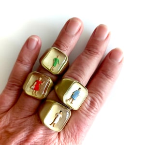 Puede incluir: Cuatro anillos dorados con caras cuadradas, cada uno con una figura en miniatura con un vestido de diferente color. Las figuras están encapsuladas en resina transparente. Los anillos se muestran en una mano sobre un fondo blanco.