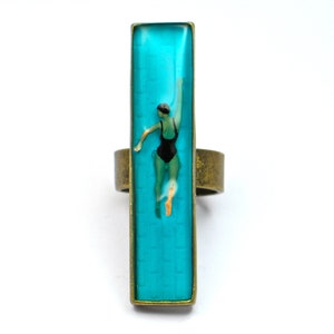 May include: A rectangular ring with a bronze band and a turquoise-colored face. The face features an image of a person in a black swimsuit swimming underwater. The ring is set against a white background.
