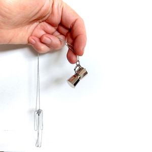 May include: A silver necklace with a small, polished metal pendant shaped like a coffee maker. The pendant is suspended from a delicate chain. The necklace is displayed against a white backdrop.