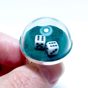 May include: A clear, dome-shaped ring with two white dice inside. The dice have black dots. The ring sits on a green surface. The ring is held by a person's hand.