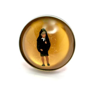 Puede incluir: Un anillo redondo, plateado, con una parte superior abovedada transparente. Dentro de la cúpula hay una figura en miniatura de una persona con abrigo y falda negros. El fondo es dorado con pequeñas motas blancas.