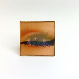 Può includere: Compact quadrato con un disegno di occhio stampato. L'immagine presenta un primo piano di un occhio con uno sfondo color pesca e una cornice dorata. L'occhio ha accenti blu, verdi e rossi.