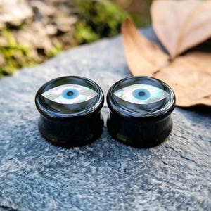 Op de afbeelding: Een paar ronde zwarte oorplugs met een ingelegd oogontwerp. Het midden van elk oog is een kleine blauwe cirkel omringd door een iriserende, parelmoerkleurige amandelvormige iris op een gladde, glanzend zwarte achtergrond.
