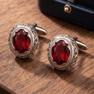 Puede incluir: Un par de gemelos plateados con una piedra preciosa de color rojo intenso de corte ovalado en el centro. El engaste de metal tiene un borde de filigrana detallado y un borde de cuentas. Se muestran con una caja azul oscuro.