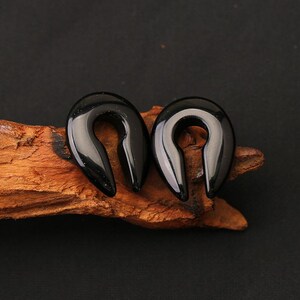 May include: A pair of black, horseshoe-shaped ear gauges with a smooth, polished finish. These U-shaped body jewelry pieces feature a solid, glossy surface and a curved design, crafted for ear stretching and body modification purposes.