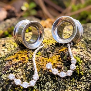 May include: A pair of silver-toned metal ear tunnels with a smooth, circular interior. Each tunnel features a dangling, curved attachment with a textured, branch-like surface adorned with five small, clear, round-cut stones set in a row.