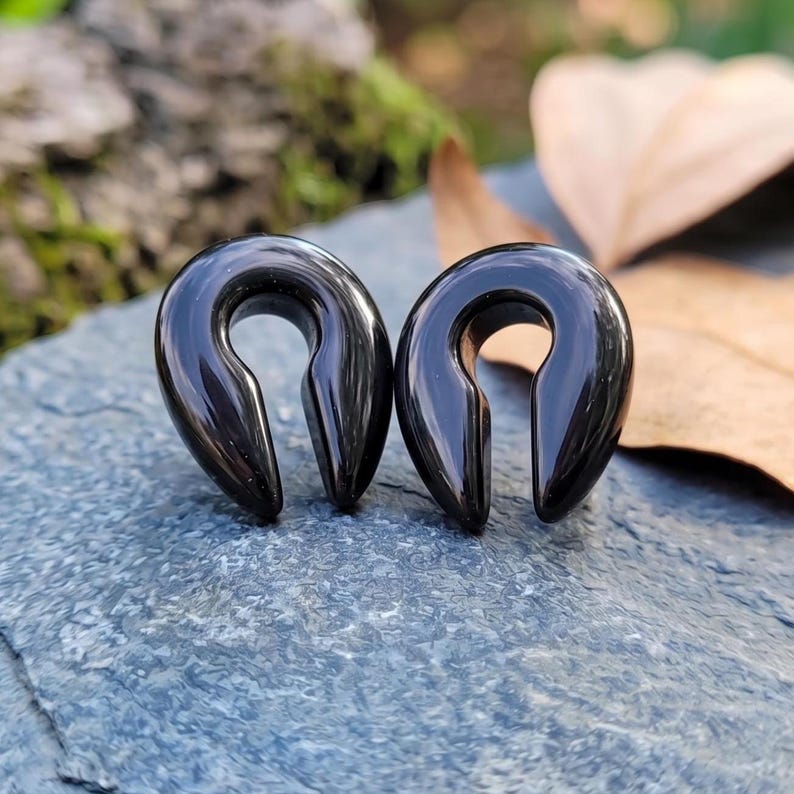 May include: A pair of black ear gauges with a smooth, glossy finish and a teardrop-shaped design. These jewelry pieces feature an open center and a tapered bottom, crafted from a solid, dark material with a reflective surface for ear stretching.