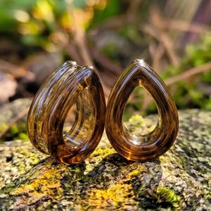 Peut inclure: Une paire d'écarteurs d'oreilles en verre translucide de couleur ambre en forme de goutte d'eau. Les écarteurs présentent une surface lisse et polie avec un centre creux, révélant une teinte brunâtre chaude qui capte la lumière.