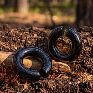 Puede incluir: Un par de pesas para orejas circulares, negras y pulidas, con un acabado liso y brillante. Cada pieza presenta una abertura distintiva, creando un diseño en forma de C. La joya tiene un perfil redondeado y grueso con una superficie reflectante.