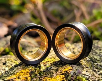 Puede incluir: Un par de dilatadores circulares con un borde exterior negro mate liso y un revestimiento interior de metal dorado pulido. Las piezas de joyería cilíndricas son huecas en el centro, mostrando un contraste marcado entre el exterior oscuro y el acabado interior dorado, reflectante y de tonos cálidos.